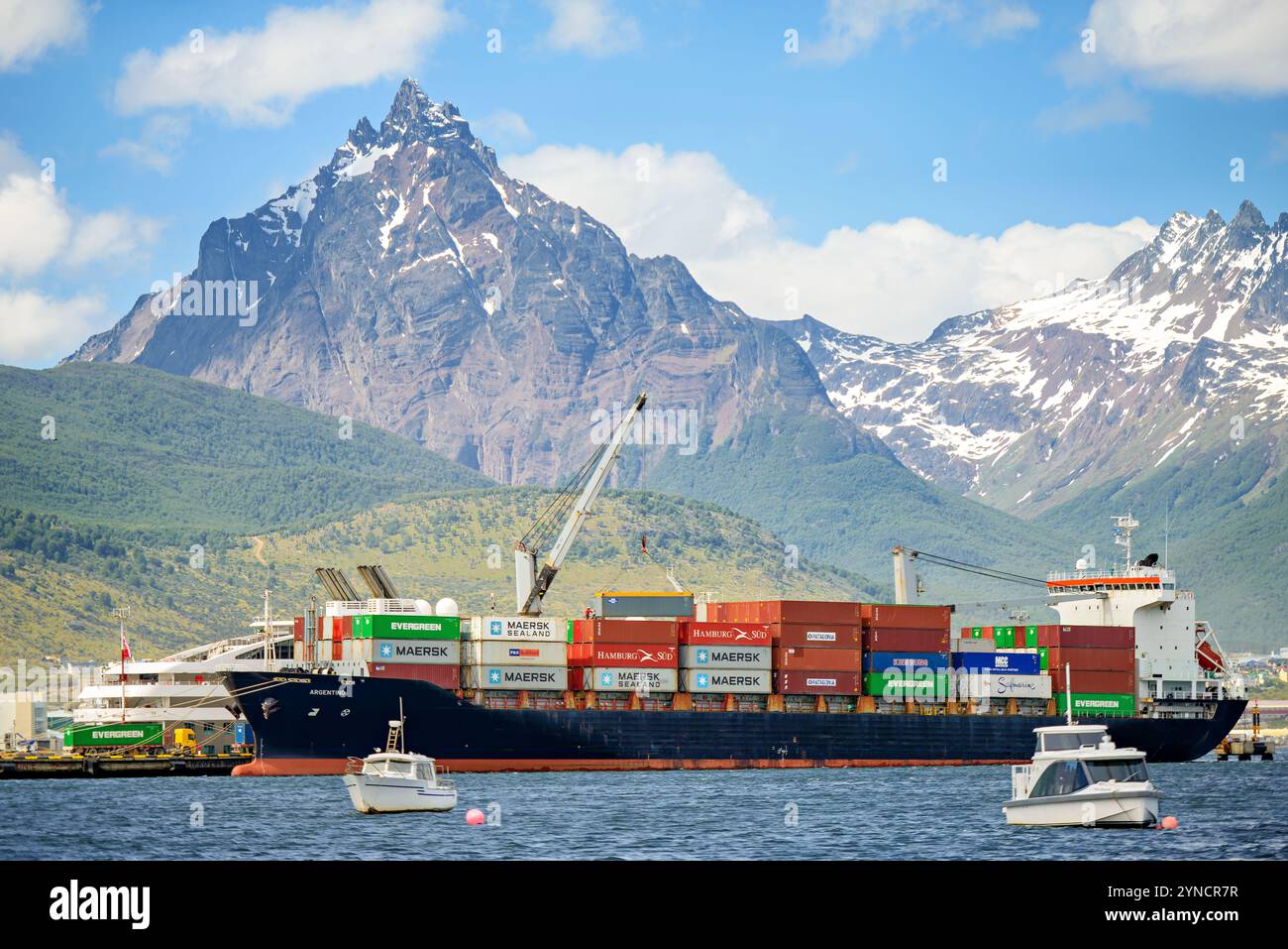 Nave portacontainer Monte Olivia Ushuaia Argentina // USHUAIA, Argentina — Una nave portacontainer attraccata al terminal merci di Ushuaia carica i container di spedizione con Monte Olivia che sale sullo sfondo. Il porto funge da centro marittimo cruciale per le operazioni di rifornimento della Terra del fuoco e dell'Antartide. Il Monte Olivia, parte delle Ande Fuegiane, raggiunge un'altitudine di 4.469 piedi (1.362 metri). Foto Stock
