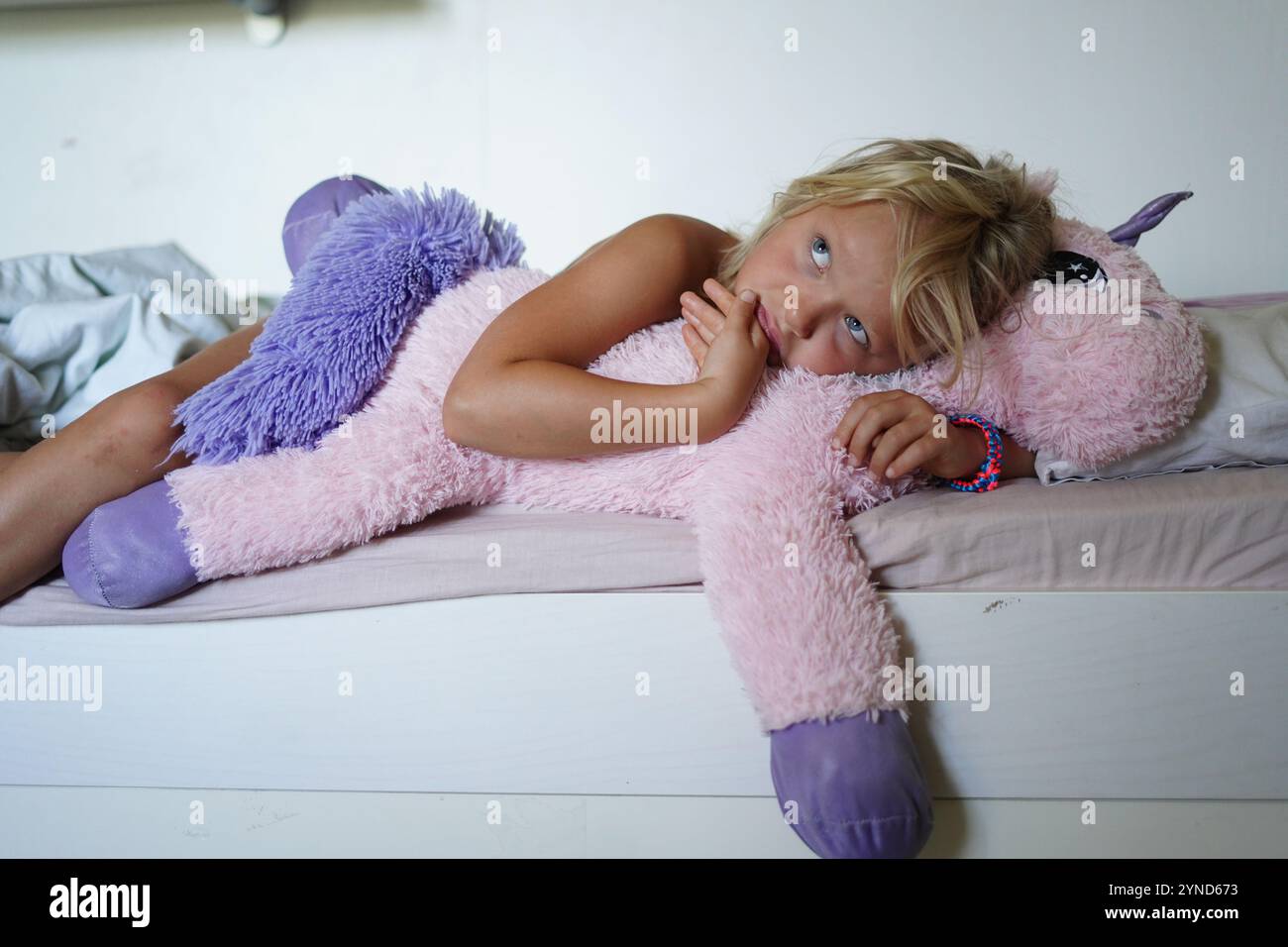 Una giovane ragazza giace a letto, sognando magiche avventure mentre coccola il suo grande unicorno rosa. L'illuminazione soffusa e l'espressione pacifica creano una vera e propria atmosfera Foto Stock