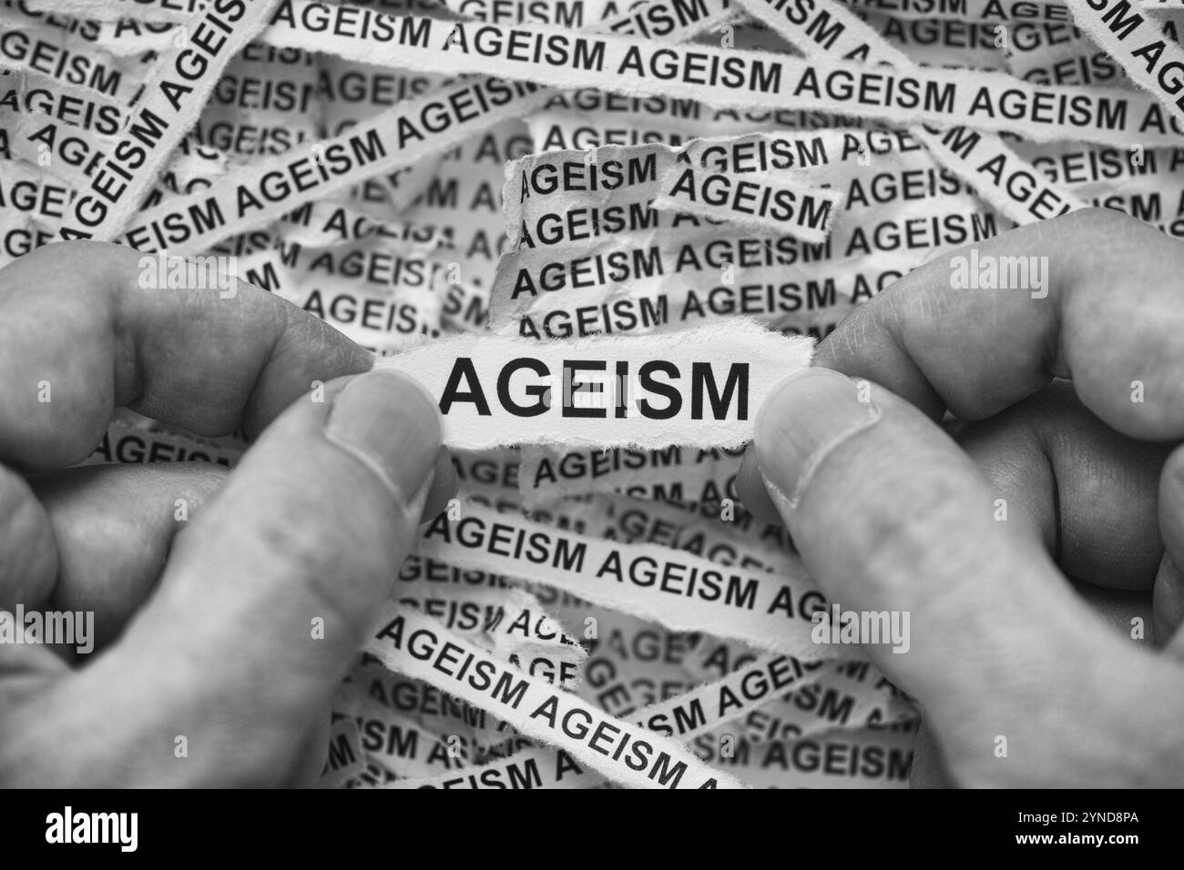Un uomo che tiene un pezzo di carta con la parola ageism tra le mani su strisce di giornale sgualcite. Bianco e nero. Primo piano. Foto Stock