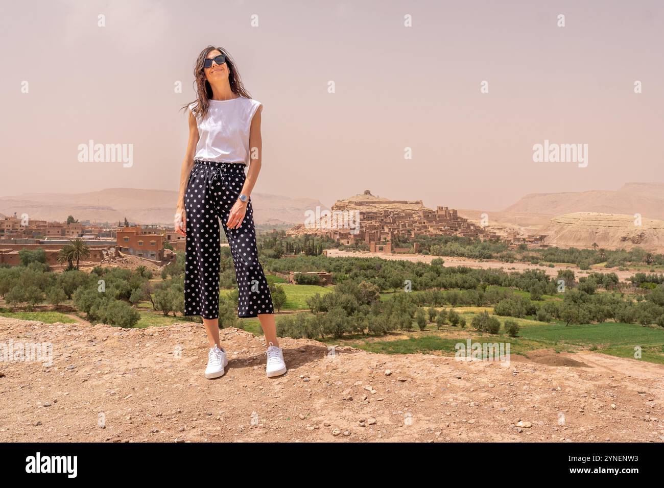 Giovane donna dai capelli scuri in pantaloni neri e camicia bianca in posa da Ait Ben Haddou Fortress, Marocco Foto Stock
