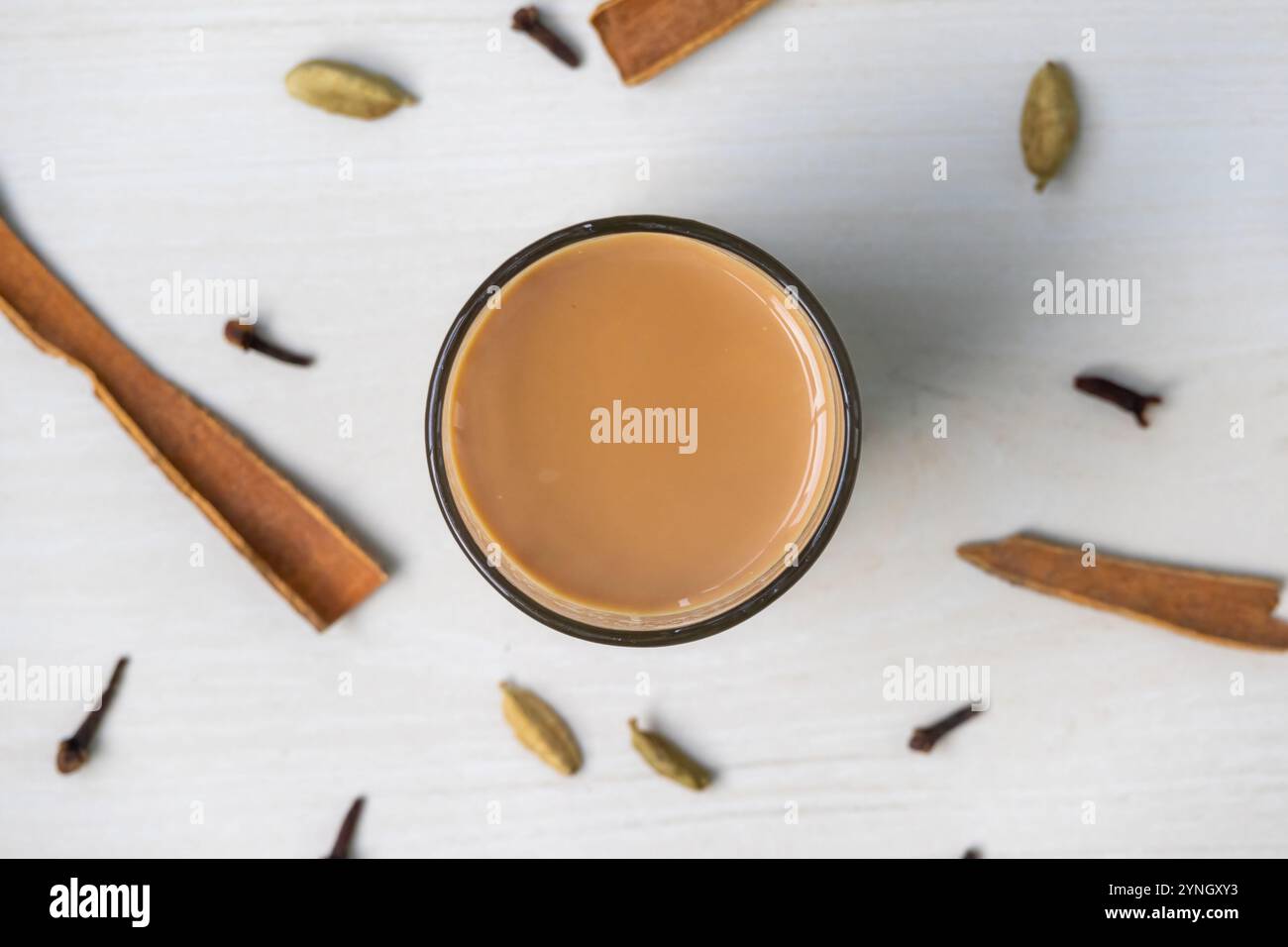 Vista dall'alto del masala chai su una tazza di vetro su una superficie di legno chiaro. Bastoncini di cannella, cardamomo e chiodi di garofano aggiungono un tocco di spezia alla scena. Foto Stock