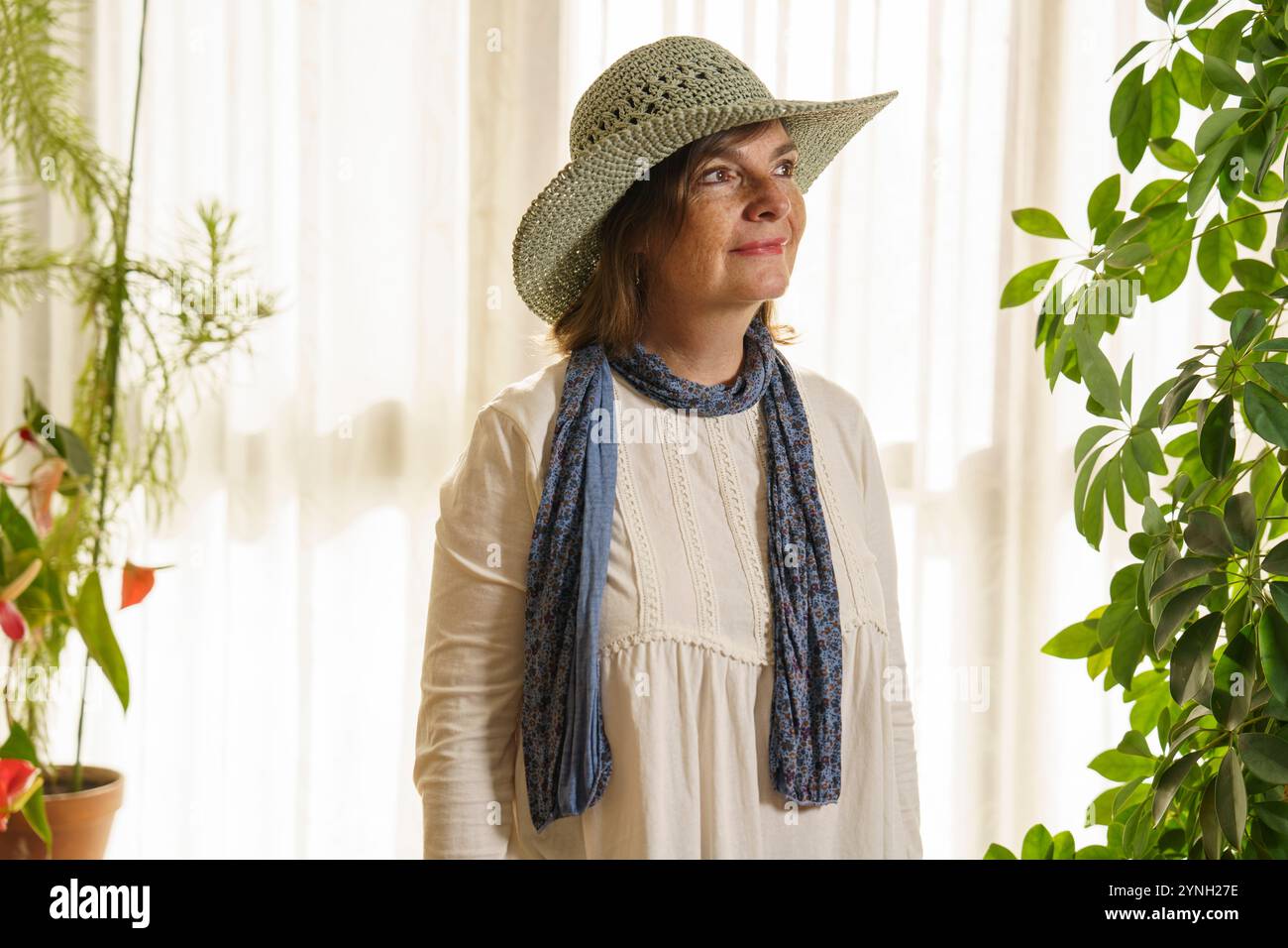 Una donna che indossa un cappello di paglia a tesa larga e una sciarpa blu si trova in una stanza illuminata dal sole piena di piante. Ha un'espressione serena, con un morbido e naturale Foto Stock