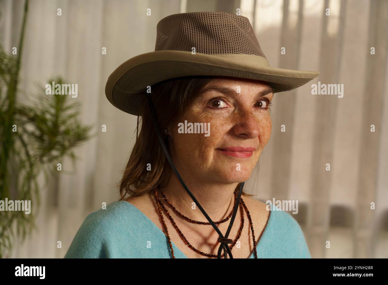 Una donna con i capelli a spalla che indossa un cappello di colore chiaro e un maglione blu. Ha un'espressione amichevole ed è in piedi in casa con un morbido natu Foto Stock Una donna con i capelli a spalla che indossa un cappello di colore chiaro e un maglione blu. Ha un'espressione amichevole ed è in piedi in casa con un morbido natu Foto Stock
