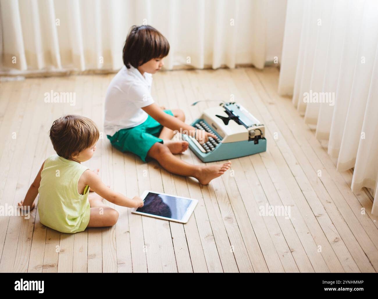 Due bambini siedono su un pavimento di legno in una stanza ben illuminata. Un bambino digita su una macchina da scrivere d'epoca, mentre l'altro interagisce con un tablet, mostrando una B. Foto Stock