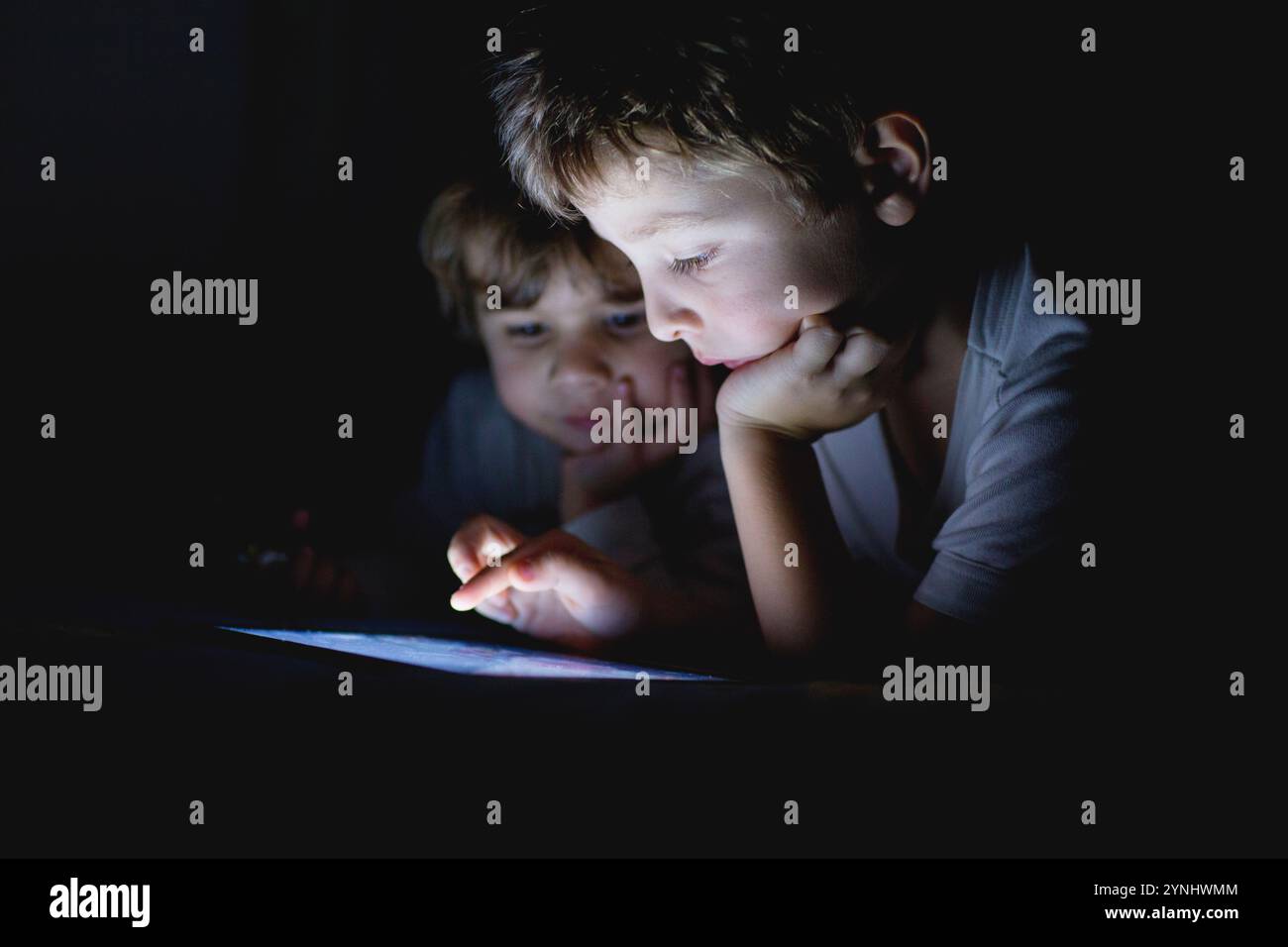 Due bambini piccoli sono impegnati con un tablet digitale in una stanza buia. Un bambino è concentrato sullo schermo, mentre l'altro guarda con curiosità, creato Foto Stock