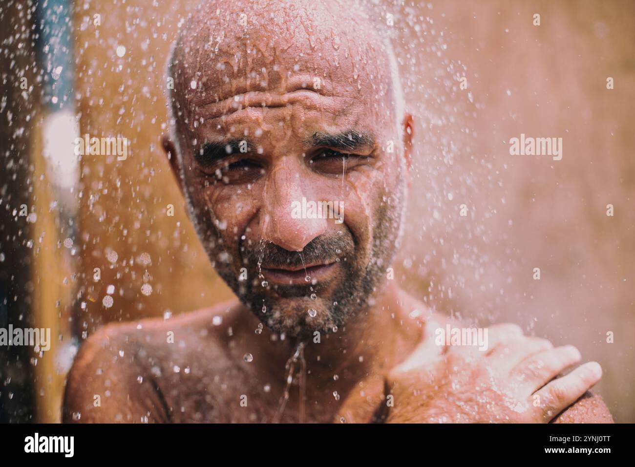 Un uomo si trova all'aperto sotto una doccia rinfrescante, con gocce d'acqua che cadono sulla pelle bagnata. Sembra rilassato, godendo del calore del sole e del Th Foto Stock