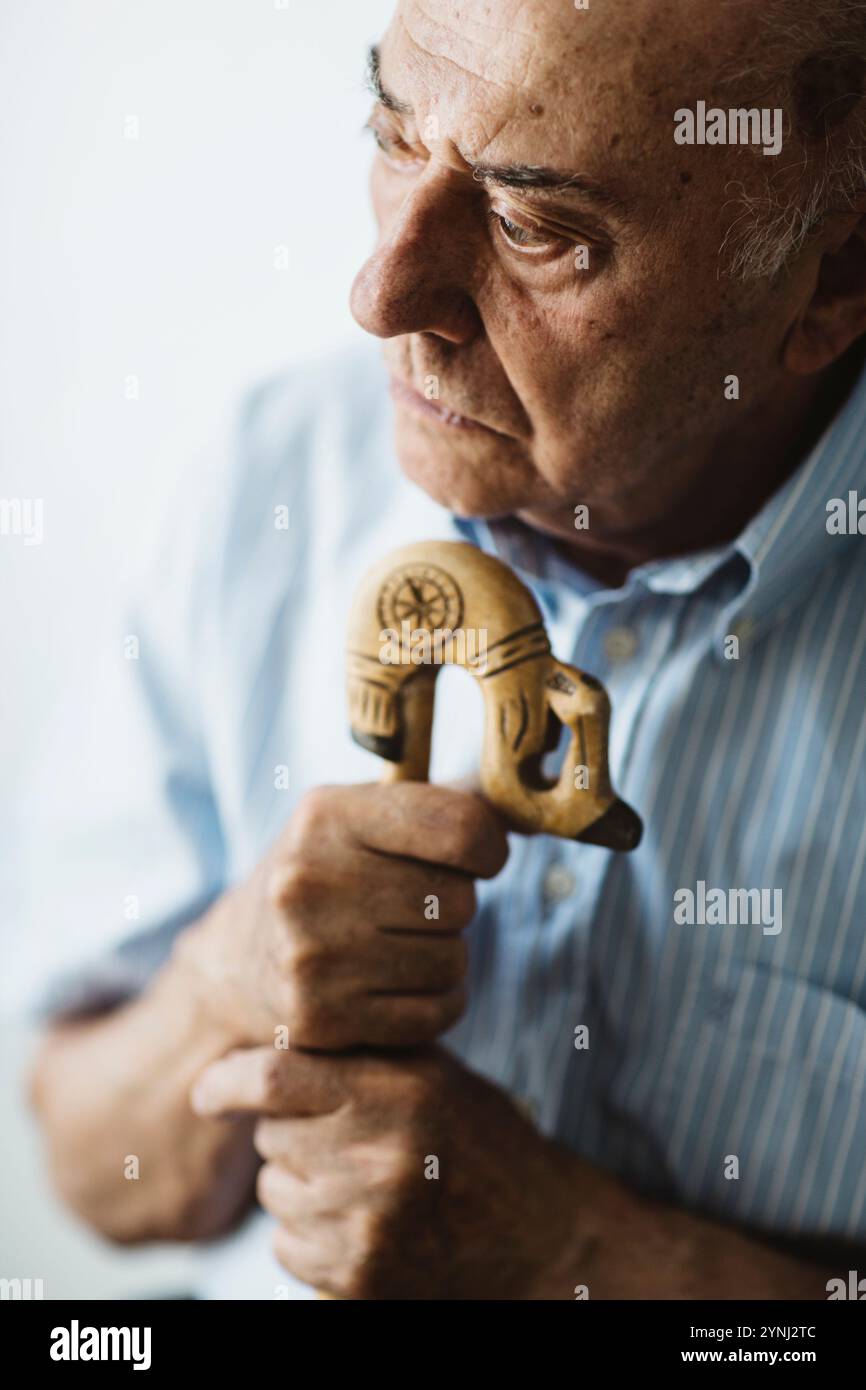 Un uomo anziano regge un bastone decorativo in legno, perso nel pensiero. Guarda verso il basso con un'espressione seria, riflettendo le complessità della vita in un tranquillo roo Foto Stock