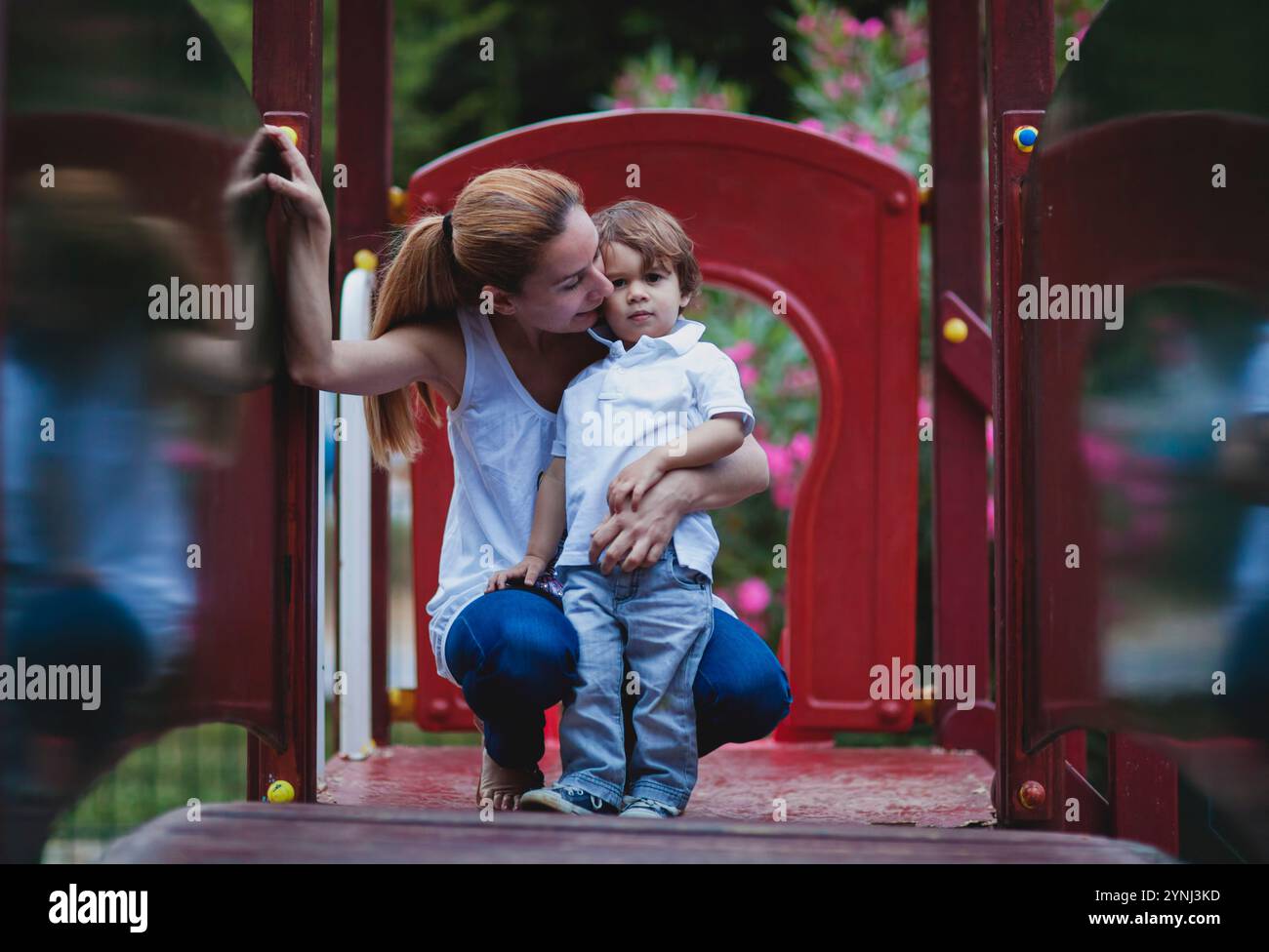 Una madre si inginocchia accanto a suo figlio, offrendo affetto mentre si rilassano sulle attrezzature del parco giochi. I vivaci fiori del parco creano uno sfondo allegro per te Foto Stock