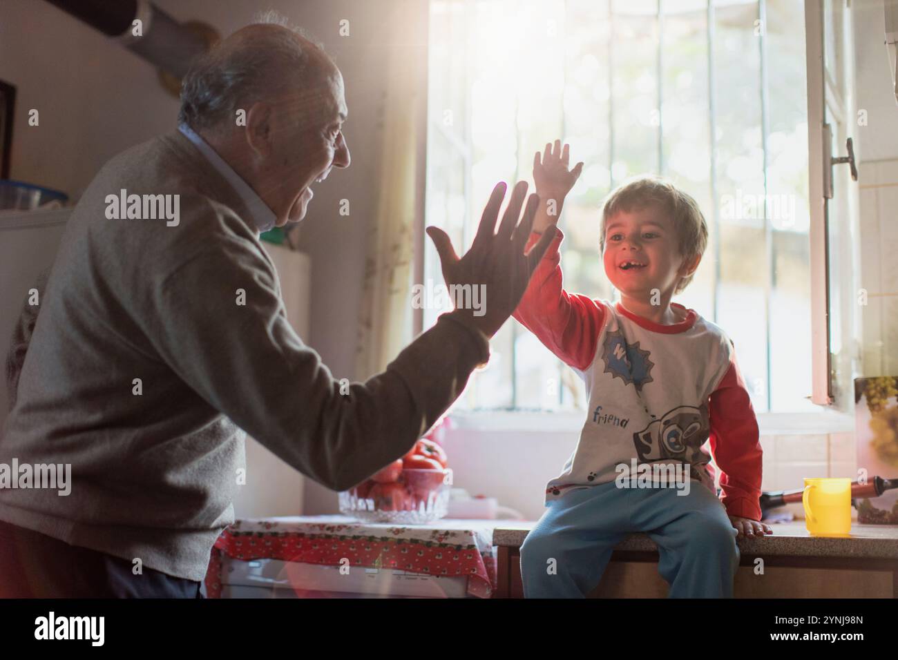 Un bambino e un anziano si impegnano in un giocoso gioco a mano vicino a una finestra. La calda luce del sole esalta le loro espressioni gioiose, creando un'atmosfera vivace Foto Stock