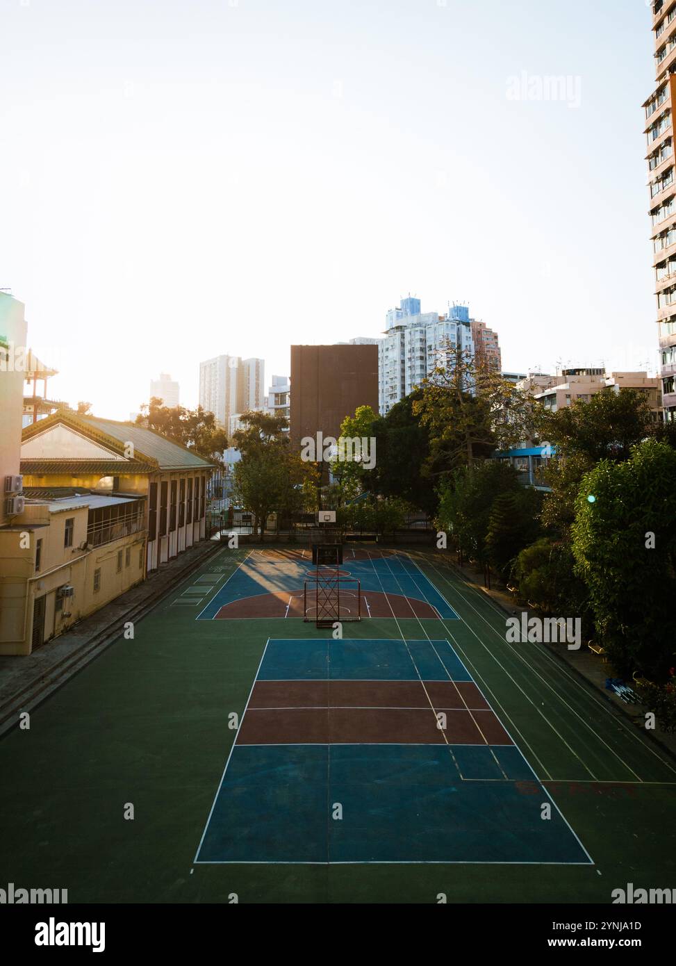 Campo da pallacanestro urbano illuminato dal sole circondato da edifici cittadini e vegetazione in un tranquillo ambiente di quartiere Foto Stock