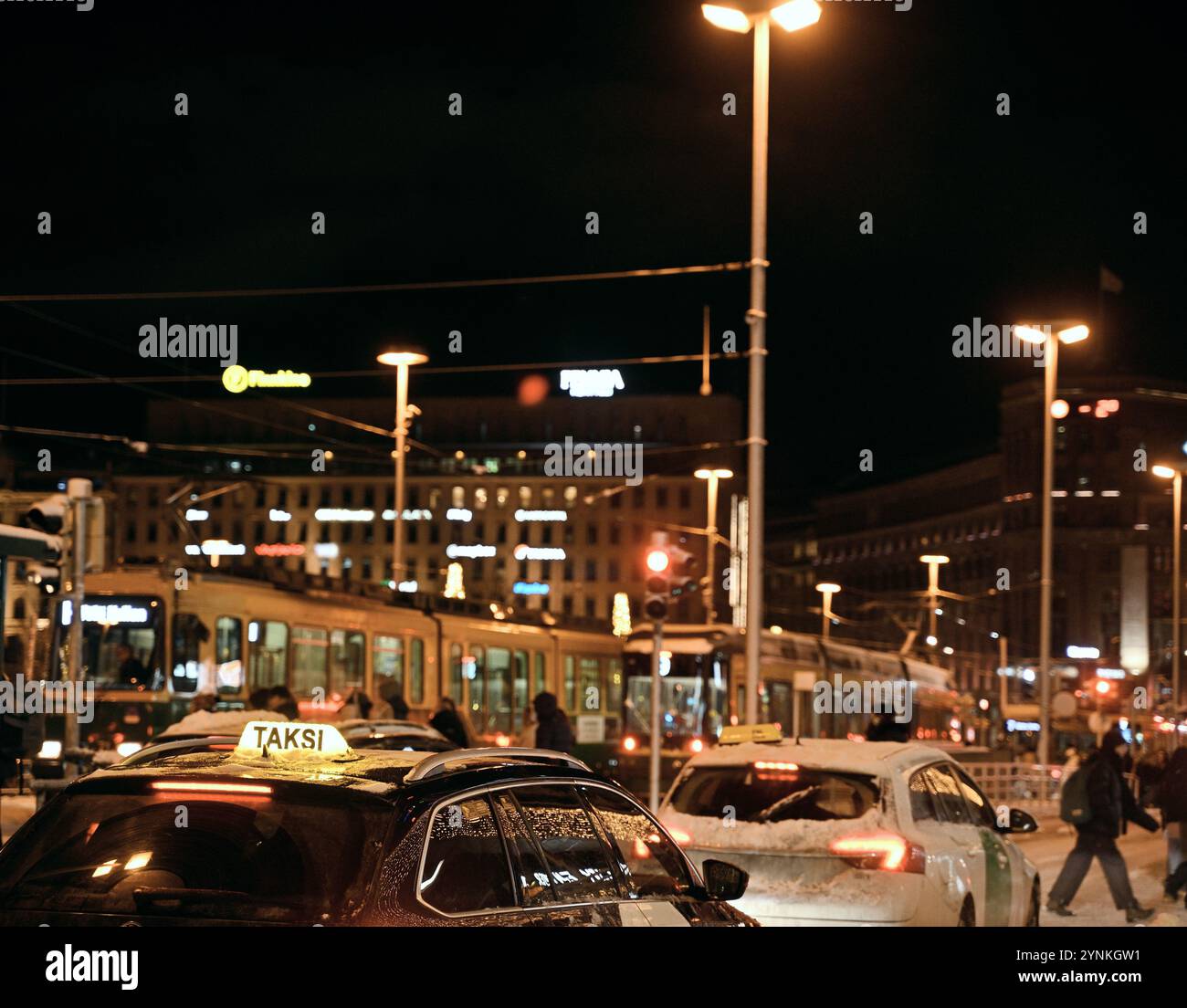 Una vivace scena notturna urbana con particolare attenzione ai taxi (cartelli "Taksi") che aspettano sotto i lampioni. Sullo sfondo è visibile un tram circondato da luce Foto Stock