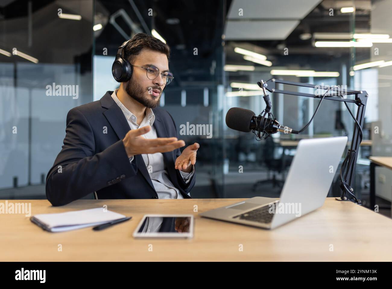 Uomo in tuta con le cuffie parla al microfono durante il podcast in ufficio. Utilizza notebook e tablet mentre comunica in modo efficace. Concetto di tecno Foto Stock