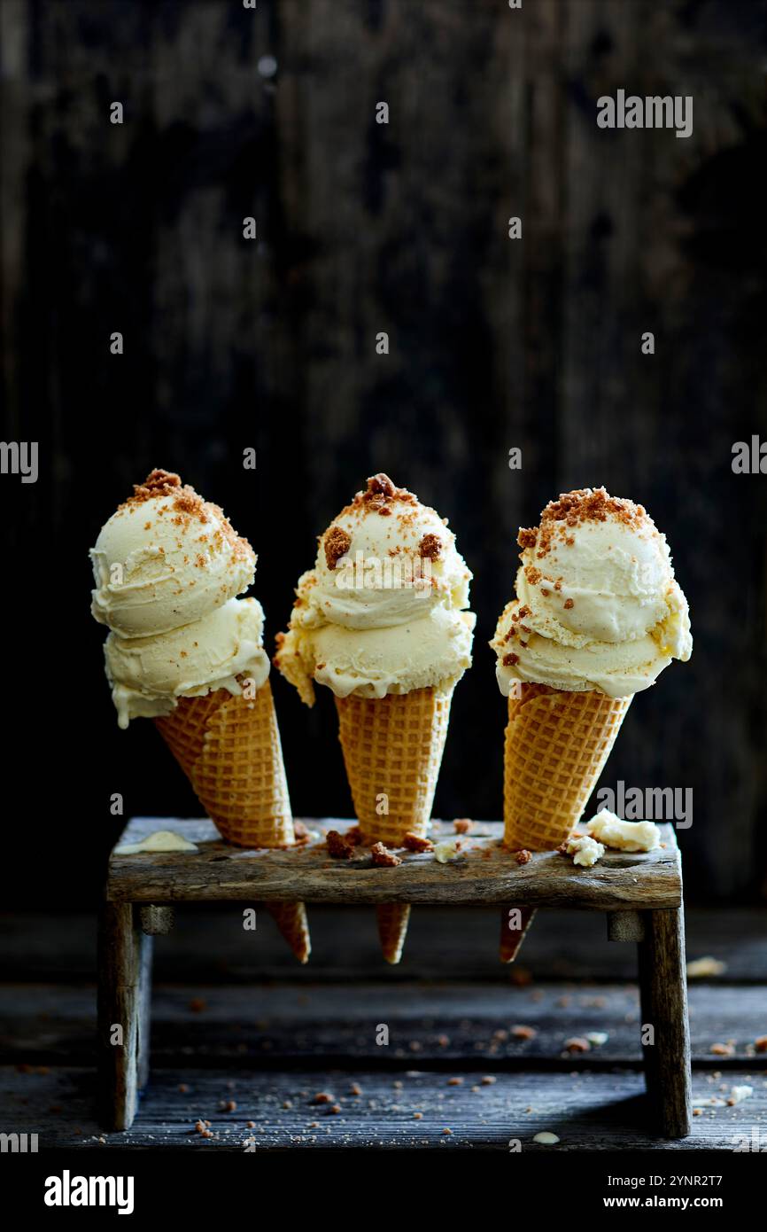 tre coni di gelato ripieni di gelato alla vaniglia e briciole di biscotti, in un banco di gelato di legno, su uno sfondo di legno scuro Foto Stock