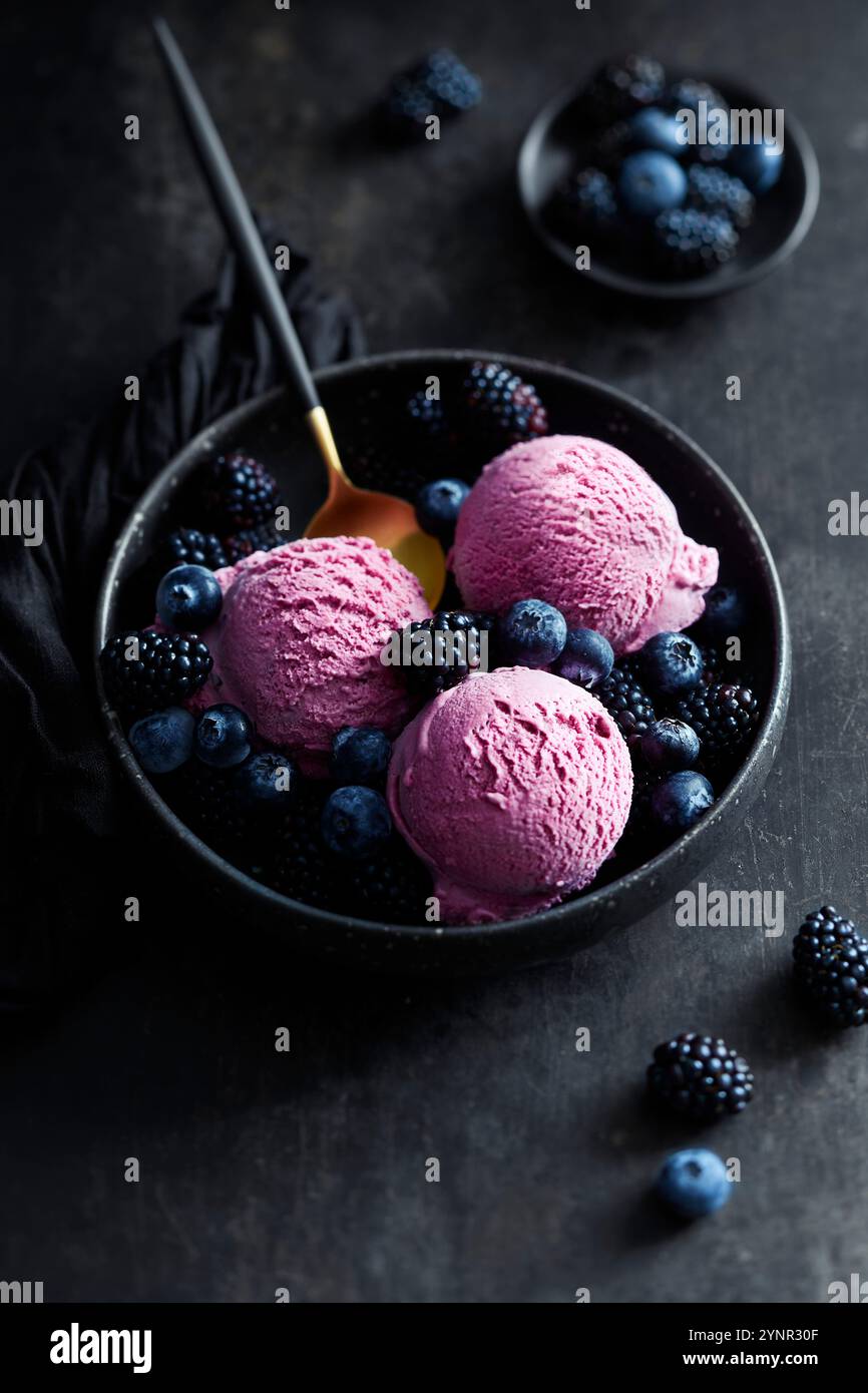 Tre palline di gelato alla mora in una ciotola nera con mirtilli, more e un cucchiaio dorato disposte su una superficie nera Foto Stock