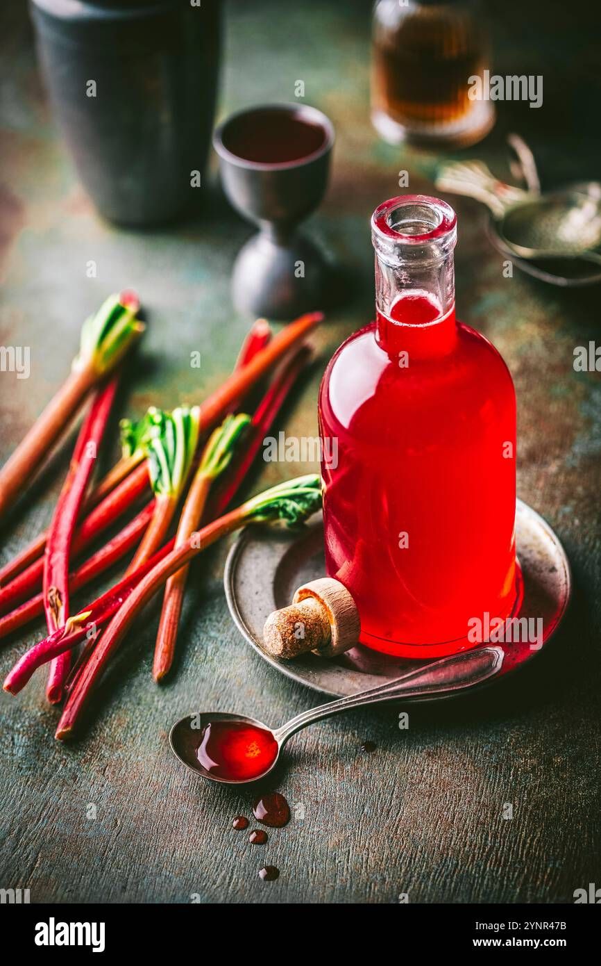 Splendida bottiglia di sciroppo di rabarbaro rosso cremisi e sciroppo in un cucchiaio, con gambi freschi di rabarbaro e cocktail jigger sullo sfondo Foto Stock