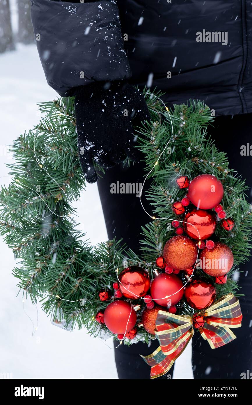 Donna irriconoscibile che regge una corona di Natale fatta a mano con decorazioni rosse. Preparazione per le festività all'aperto in inverno con il clima innevato. Creazione di regali per le vacanze sostenibili e responsabili. Copia spazio Foto Stock