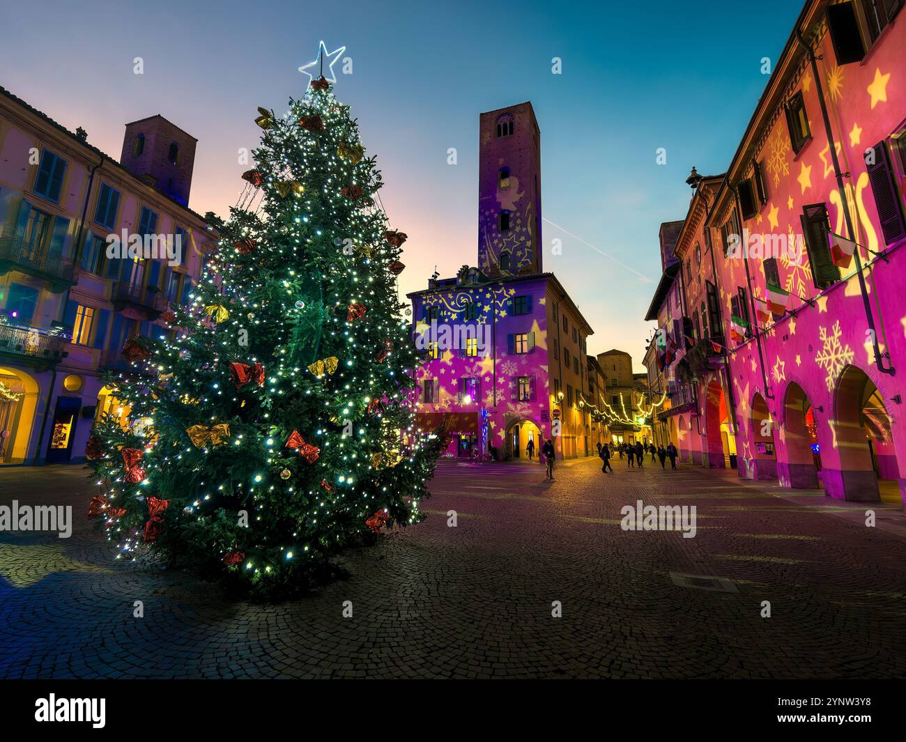 Albero di Natale illuminato al centro di una piazza acciottolata al crepuscolo circondata da edifici storici con proiezioni colorate sulle pareti. Foto Stock