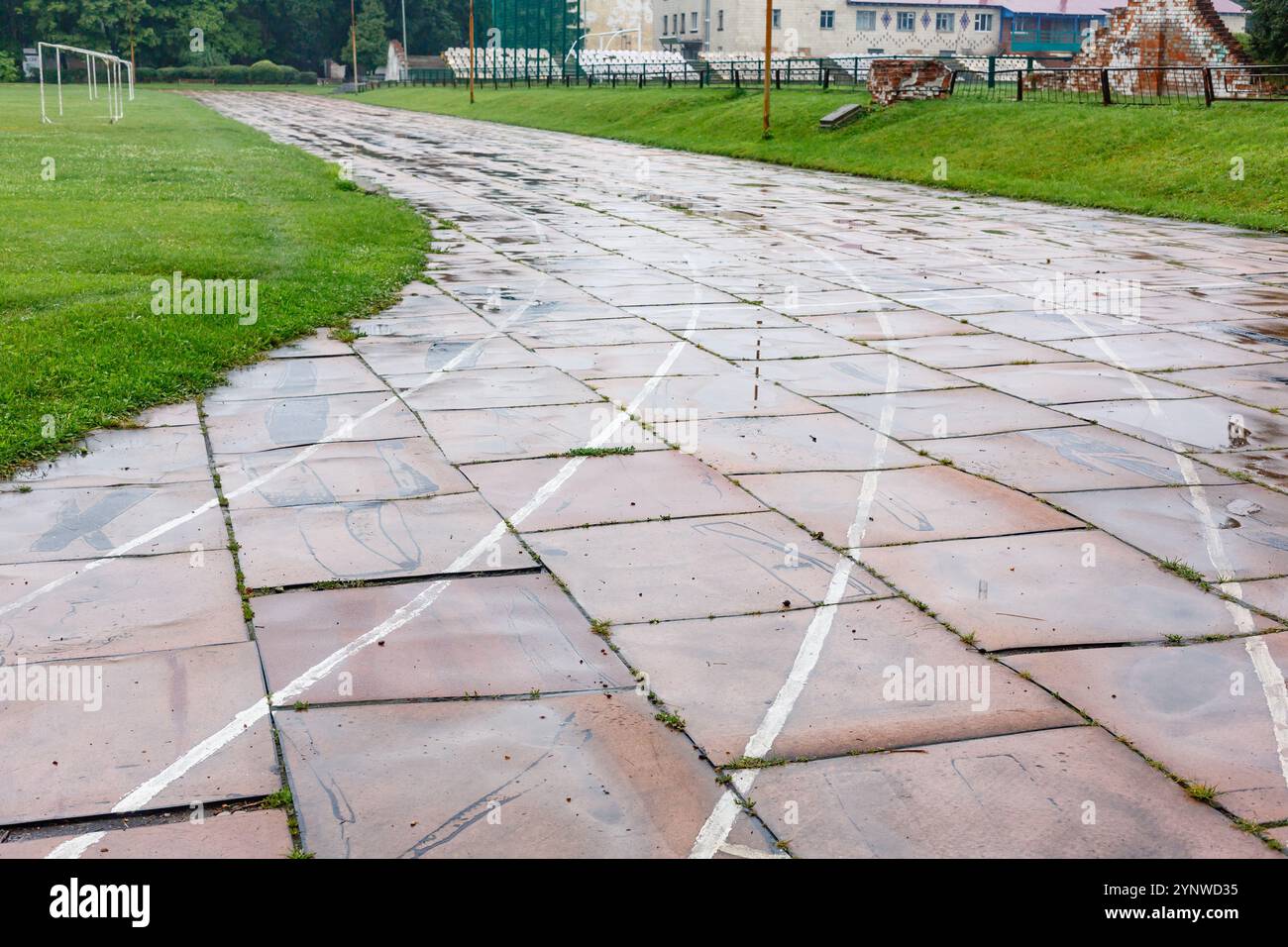 Vecchia pista da corsa danneggiata nello stadio con linee bianche sbiadite e piastrelle rotte, circondata da erba verde e campi. Concetto di strutture sportive urbane. Foto Stock