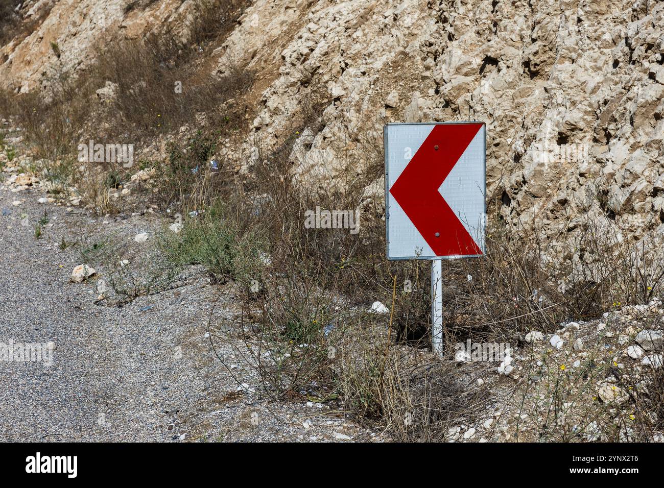 Un segnale stradale curvo rosso e bianco si trova accanto a una strada di ghiaia tra i dintorni rocciosi nell'Iran rurale, a indicare una curva stretta davanti. Foto Stock