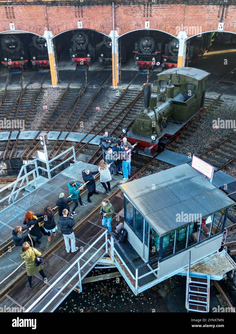 27 novembre 2024, Sassonia, Chemnitz: Con forze combinate, gli ospiti del Museo ferroviario Sassone di Chemnitz tirano fuori dal capannone la locomotiva a vapore "Hegel" (98 7056) di oltre 20 tonnellate (foto con drone). La locomotiva, costruita nel 1886 presso la Sächsische Maschinenfabrik vormals Richard Hartmann AG, è una delle mostre ed è stata ora estratta dal capannone da volontari su base di test. Per la cerimonia di apertura dell'anno della capitale della cultura, un totale di dieci squadre di 12 persone sono state ricercate per tirare la locomotiva normalmente da 250 cv a una certa distanza in una competizione. Questo evento fa parte dell'operazione Foto Stock