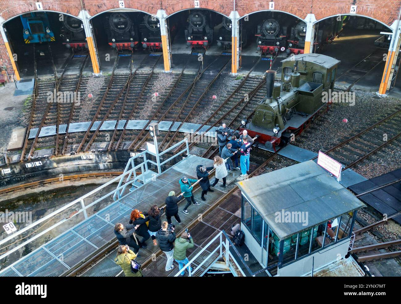 27 novembre 2024, Sassonia, Chemnitz: Con forze combinate, gli ospiti del Museo ferroviario Sassone di Chemnitz tirano fuori dal capannone la locomotiva a vapore "Hegel" (98 7056) di oltre 20 tonnellate (foto con drone). La locomotiva, costruita nel 1886 presso la Sächsische Maschinenfabrik vormals Richard Hartmann AG, è una delle mostre ed è stata ora estratta dal capannone da volontari su base di test. Per la cerimonia di apertura dell'anno della capitale della cultura, un totale di dieci squadre di 12 persone sono state ricercate per tirare la locomotiva normalmente da 250 cv a una certa distanza in una competizione. Questo evento fa parte di Th Foto Stock