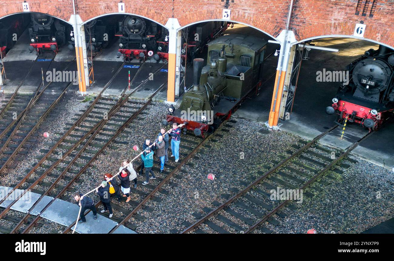 27 novembre 2024, Sassonia, Chemnitz: Con forze combinate, gli ospiti del Museo ferroviario Sassone di Chemnitz tirano fuori dal capannone la locomotiva a vapore "Hegel" (98 7056) di oltre 20 tonnellate (foto con drone). La locomotiva, costruita nel 1886 presso la Sächsische Maschinenfabrik vormals Richard Hartmann AG, è una delle mostre ed è stata ora estratta dal capannone da volontari su base di test. Per la cerimonia di apertura dell'anno della capitale della cultura, un totale di dieci squadre di 12 persone sono state ricercate per tirare la locomotiva normalmente da 250 cv a una certa distanza in una competizione. Questo evento fa parte Foto Stock