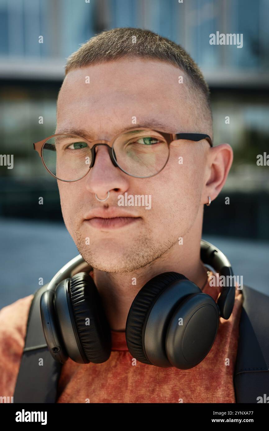 Ritratto verticale di un uomo adulto con le cuffie che indossano gli occhiali che guardano la fotocamera mentre si è in piedi fuori in città su uno sfondo sfocato, poliziotto Foto Stock