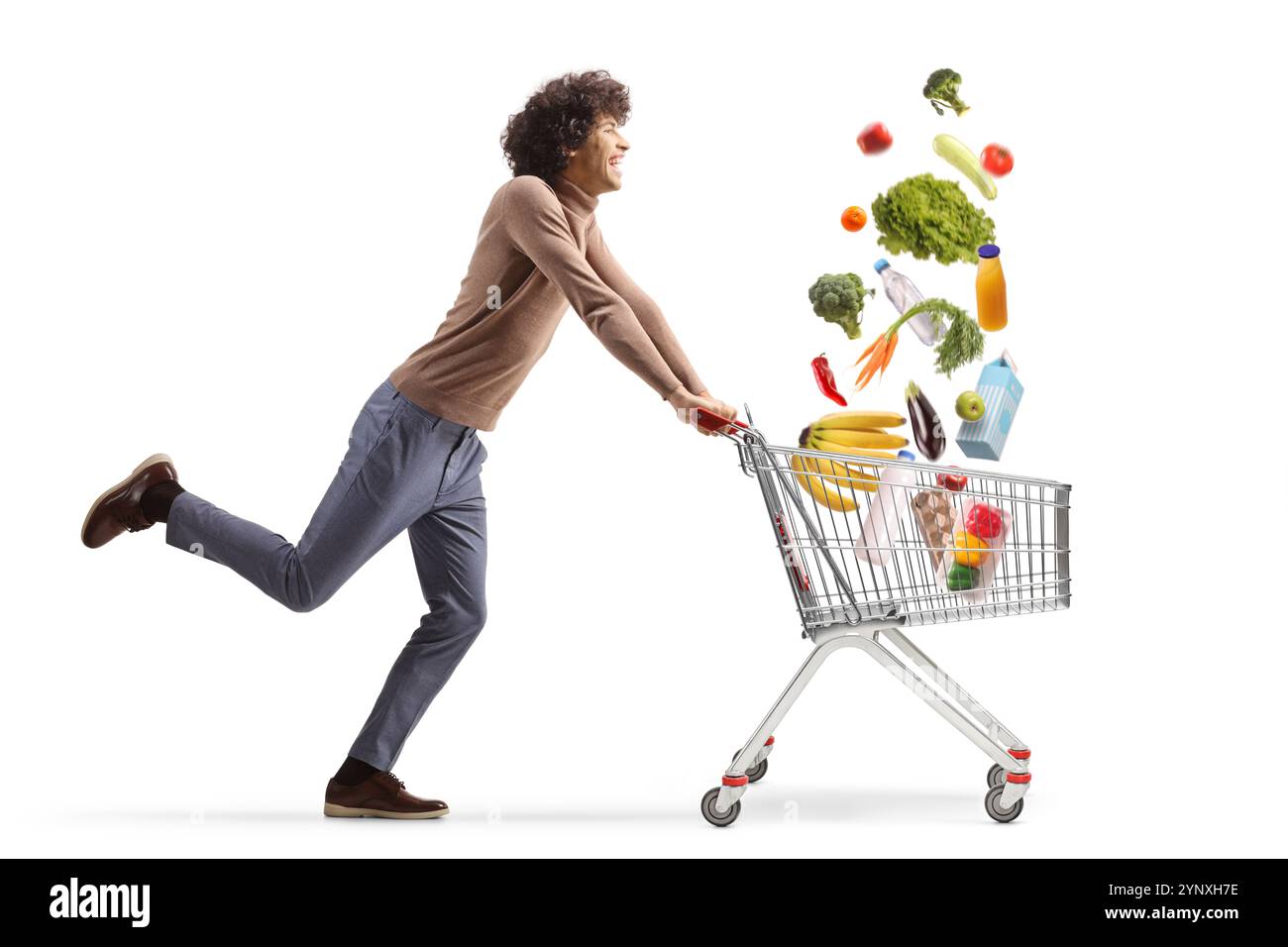 Foto a tutta lunghezza di un giovane uomo che corre con un carrello della spesa e raccoglie generi alimentari in caduta isolati su sfondo bianco Foto Stock