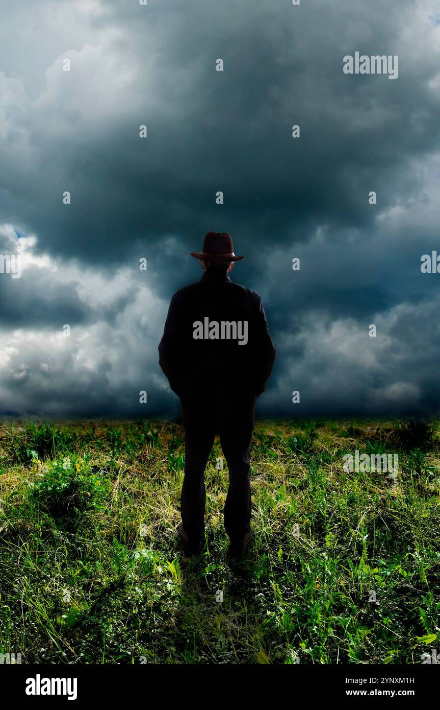 uomo che indossa un cappello fedora, in piedi in un prato, sopra un cielo tempestoso Foto Stock