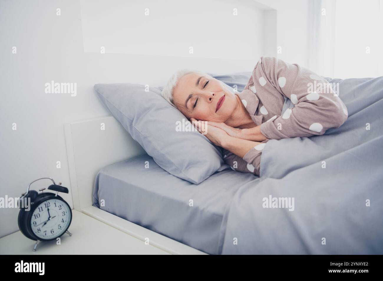 Donna anziana che dorme tranquillamente nel letto indossando pigiama a pois, abbracciando serenità e relax a casa con luce naturale Foto Stock