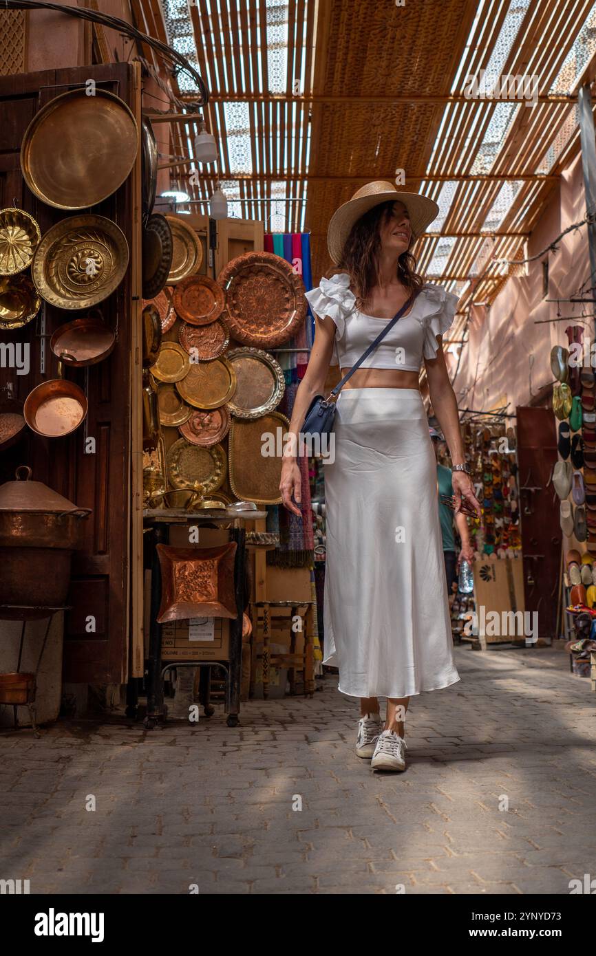 Giovane donna dai capelli scuri con abito bianco e cappello in posa in una vivace via dello shopping di Marrakech, Marocco Foto Stock