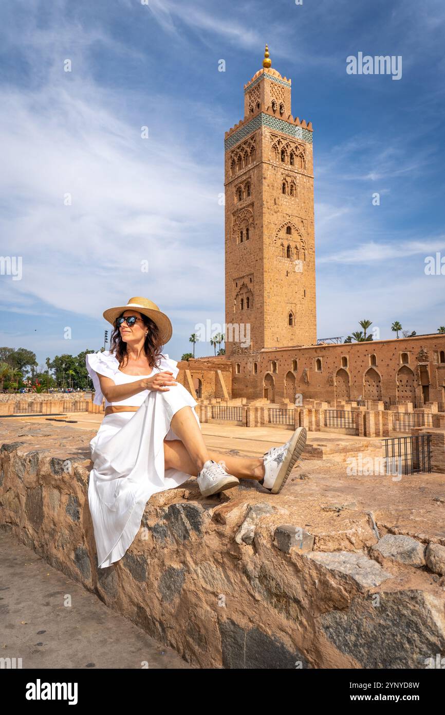 Giovane donna dai capelli scuri in abito bianco e cappello di paglia con Koutoubia Mosque Tower sullo sfondo, Marocco Foto Stock