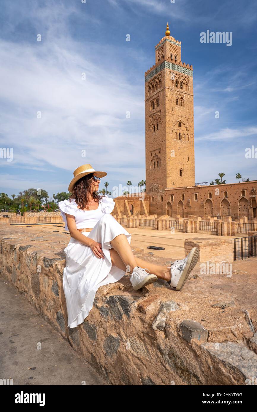 Giovane donna dai capelli scuri in abito bianco e cappello di paglia con Koutoubia Mosque Tower sullo sfondo, Marocco Foto Stock