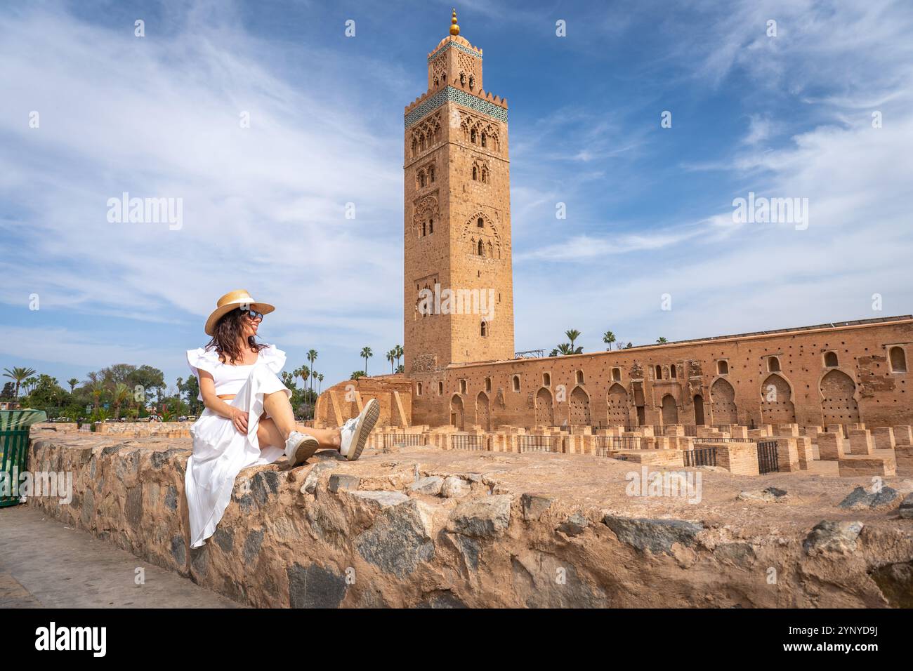 Giovane donna dai capelli scuri in abito bianco e cappello di paglia con Koutoubia Mosque Tower sullo sfondo, Marocco Foto Stock