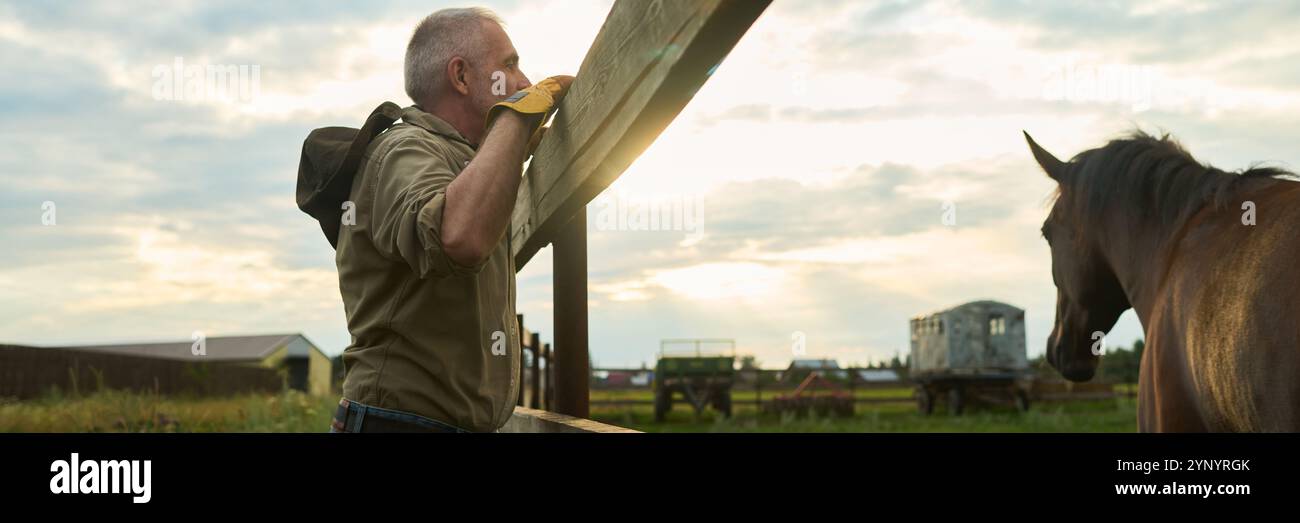 Panoramica di un uomo anziano in un abbigliamento informale in piedi accanto al recinto del bestiame con cavallo di razza marrone che pascolano sull'erba verde il giorno d'estate Foto Stock