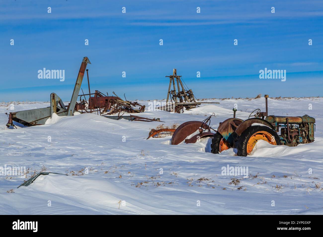 Un trattore è seduto nella neve accanto a un mucchio di vecchie attrezzature. La scena è desolata e abbandonata, con la neve che copre il terreno e la vecchia m Foto Stock