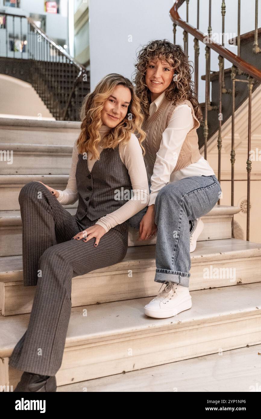 Due giovani donne d'affari sedute su gradini di marmo all'interno di un edificio, godendosi un momento di relax durante la loro giornata di lavoro Foto Stock