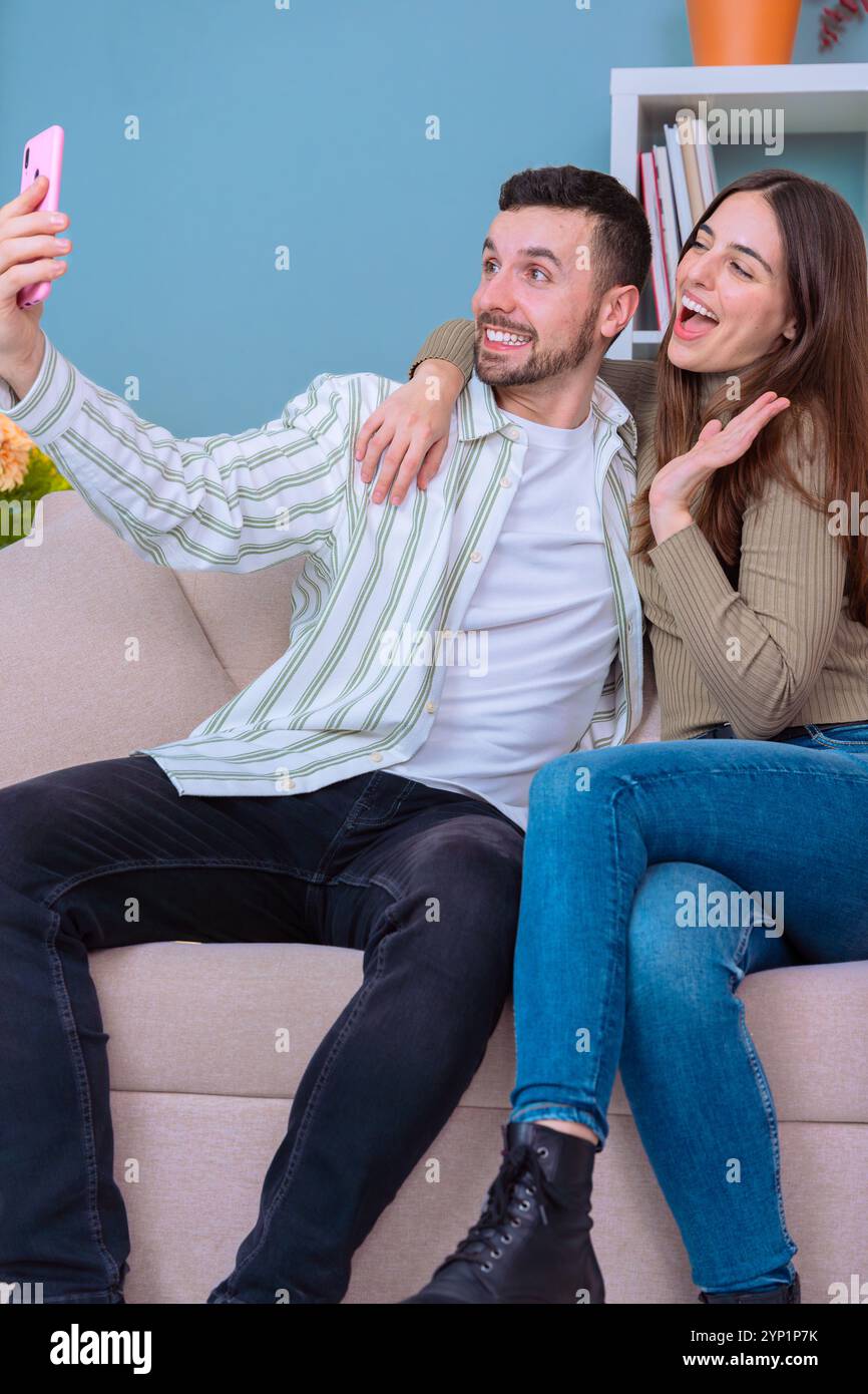 Coppia che scatta selfie sul divano nell'accogliente soggiorno con smartphone e sorride. Foto Stock