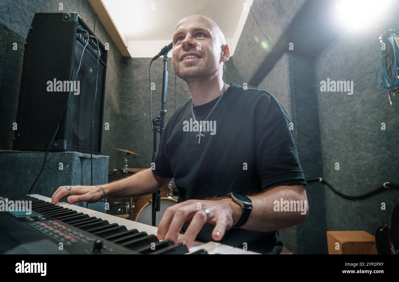 Musicista che suona la tastiera in studio di registrazione, sorridendo e godendo di un processo creativo. Attrezzatura professionale per produttori musicali in background, atmosfera stimolante e dinamica per la creatività. Foto Stock