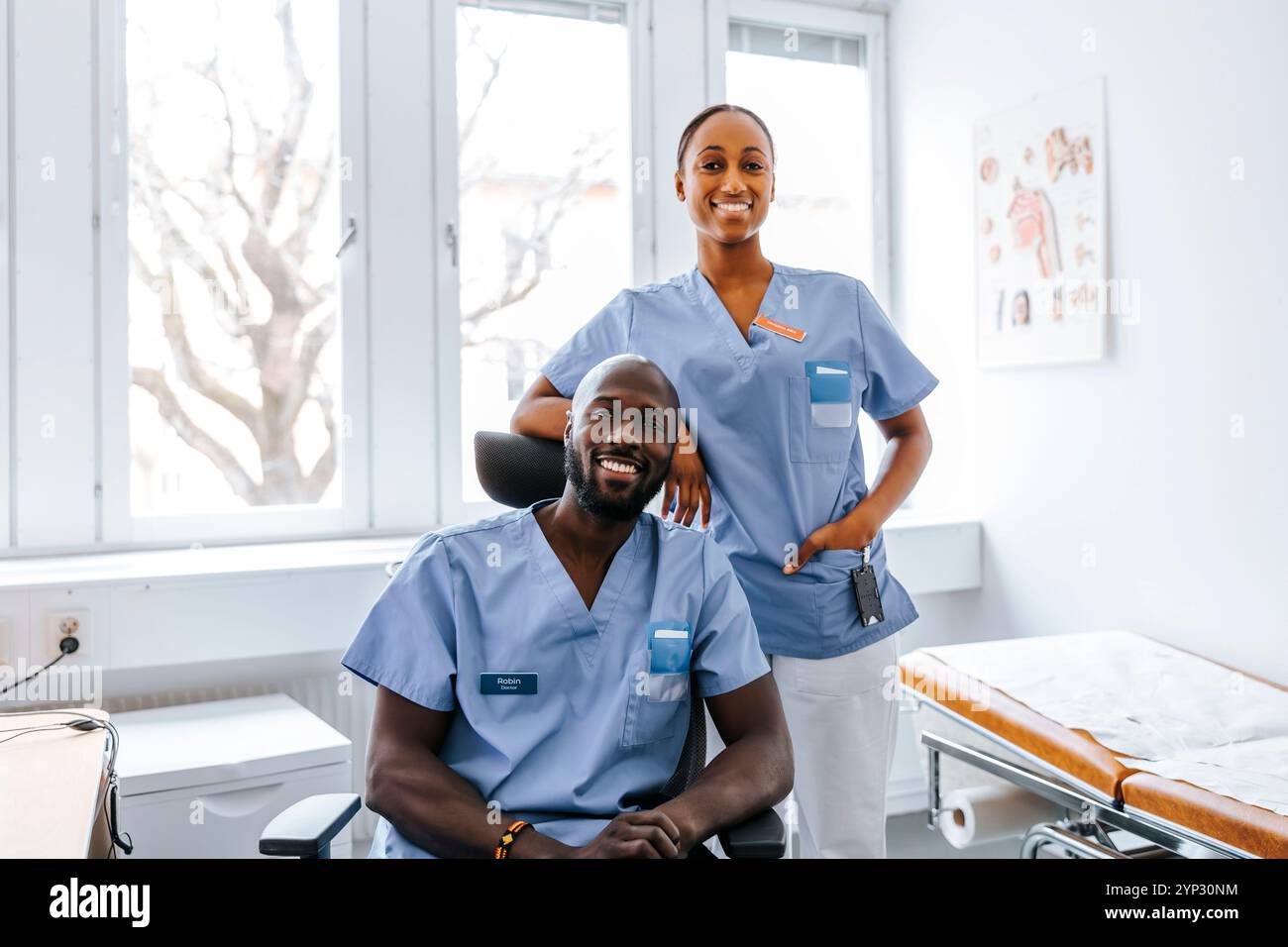 Ritratto di medico donna sorridente con mano in tasca mentre si trova accanto al collega maschio seduto sulla sedia in medico e Foto Stock