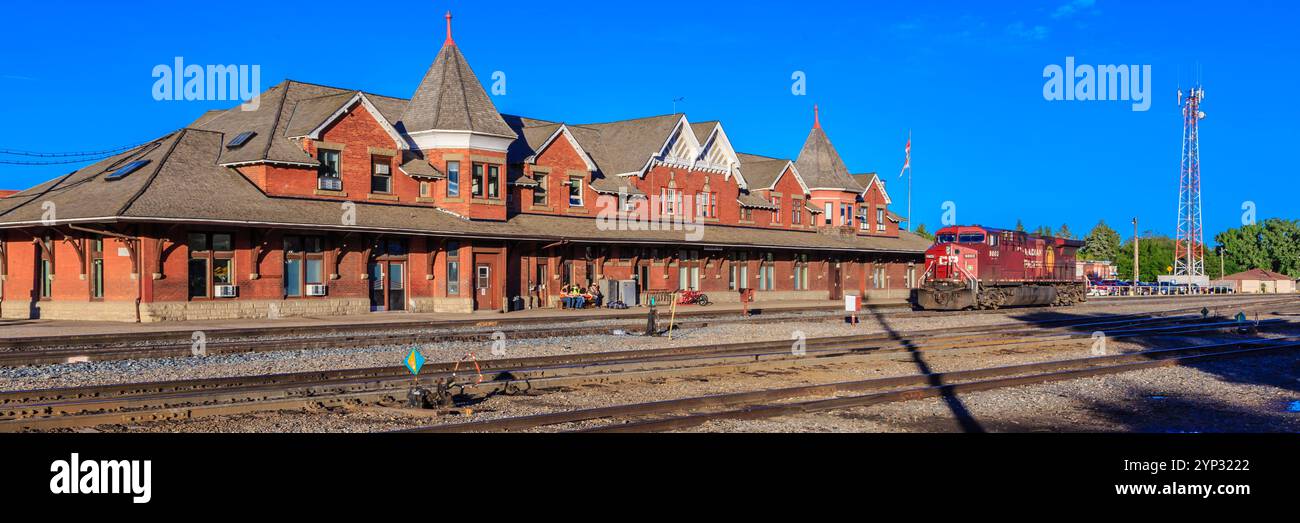 Un treno sta entrando in una stazione ferroviaria con un grande edificio rosso sullo sfondo Foto Stock