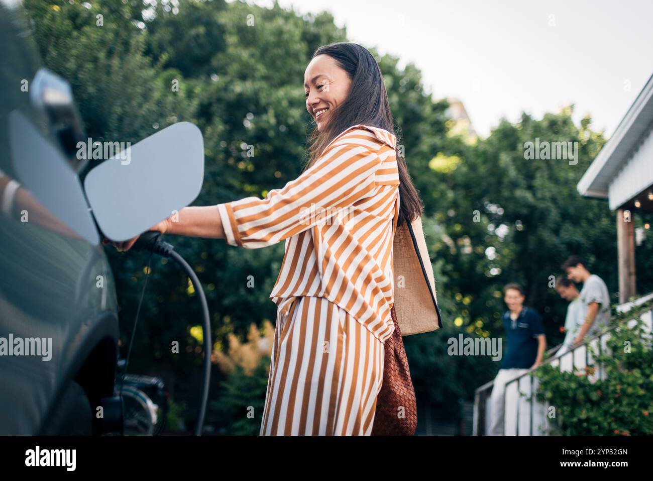 Visuale ad angolo basso della donna sorridente che carica l'auto con la spina elettrica nel cortile posteriore Foto Stock