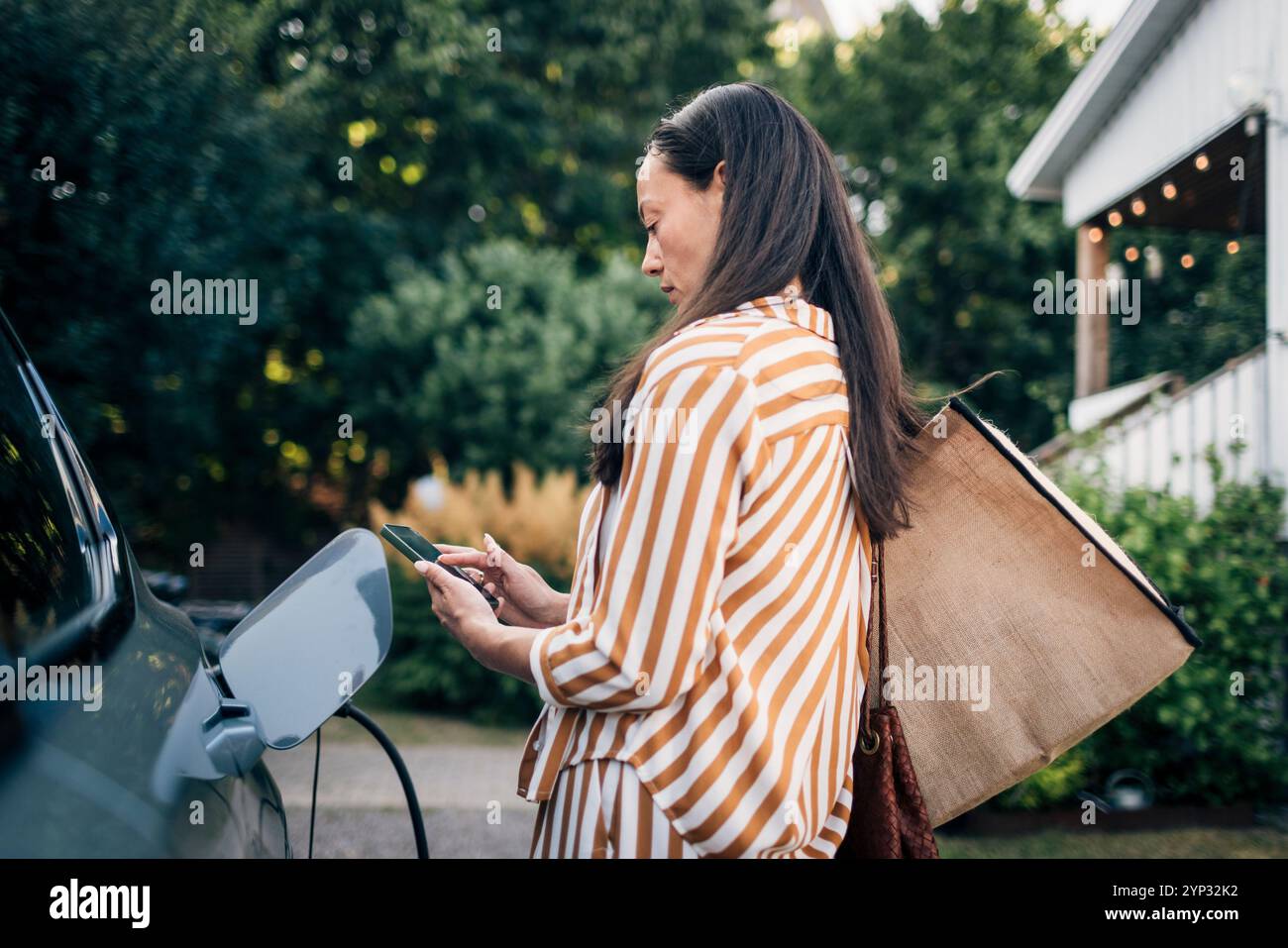 Donna concentrata che trasporta borse e utilizza lo smartphone mentre ricarica l'auto nel cortile posteriore Foto Stock