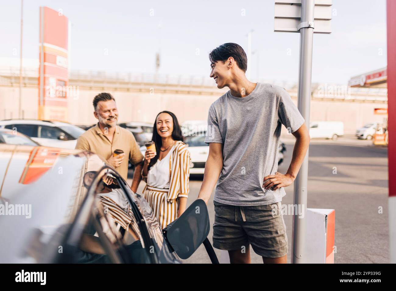 Giovane felice che ricarica l'auto con la spina elettrica mentre parla con i genitori alla stazione Foto Stock