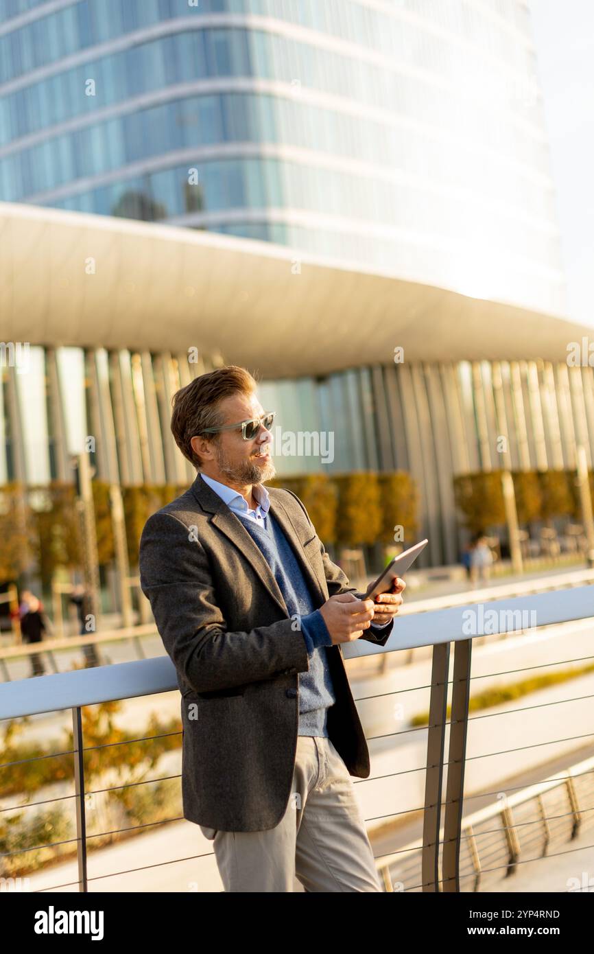 Un individuo elegante in un blazer si erge su un balcone al tramonto, tenendo in mano un tablet e guardando sopra una vivace metropoli, incarnando professionalità e. Foto Stock