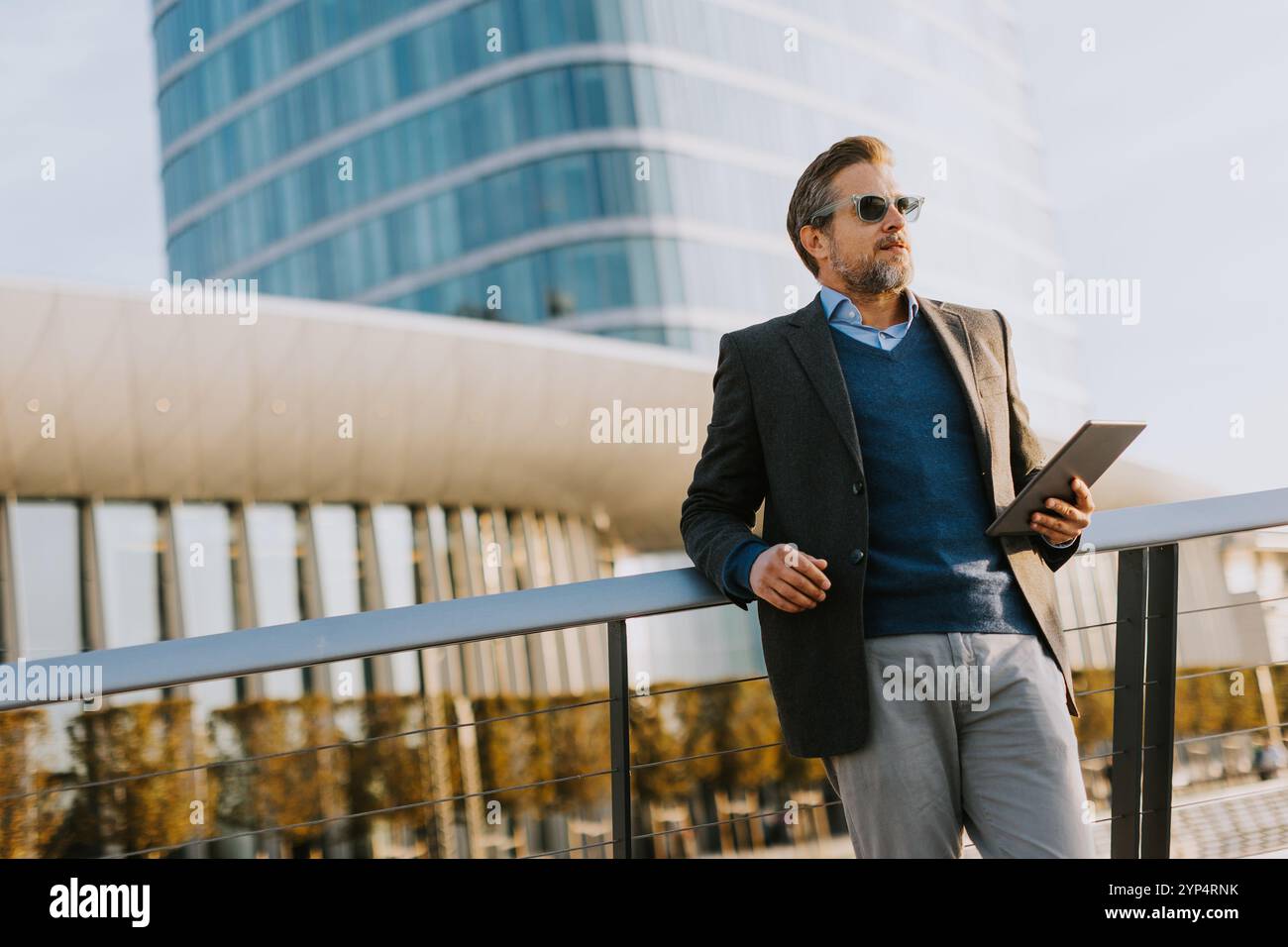 Un individuo elegante in un blazer si erge su un balcone al tramonto, tenendo in mano un tablet e guardando sopra una vivace metropoli, incarnando professionalità e. Foto Stock
