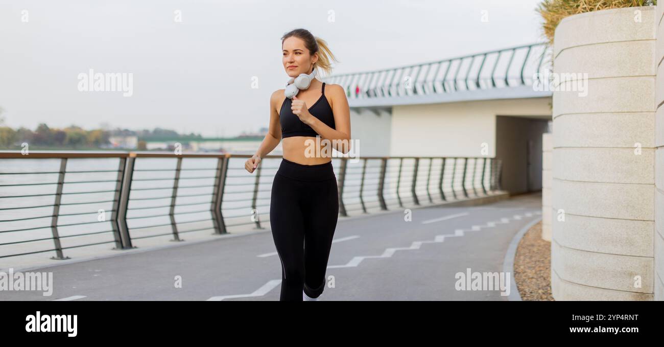 Una donna allegra corre senza sforzo lungo un pittoresco sentiero lungo il fiume, godendosi l'aria fresca e l'ambiente vivace durante una mattinata vivace. Foto Stock