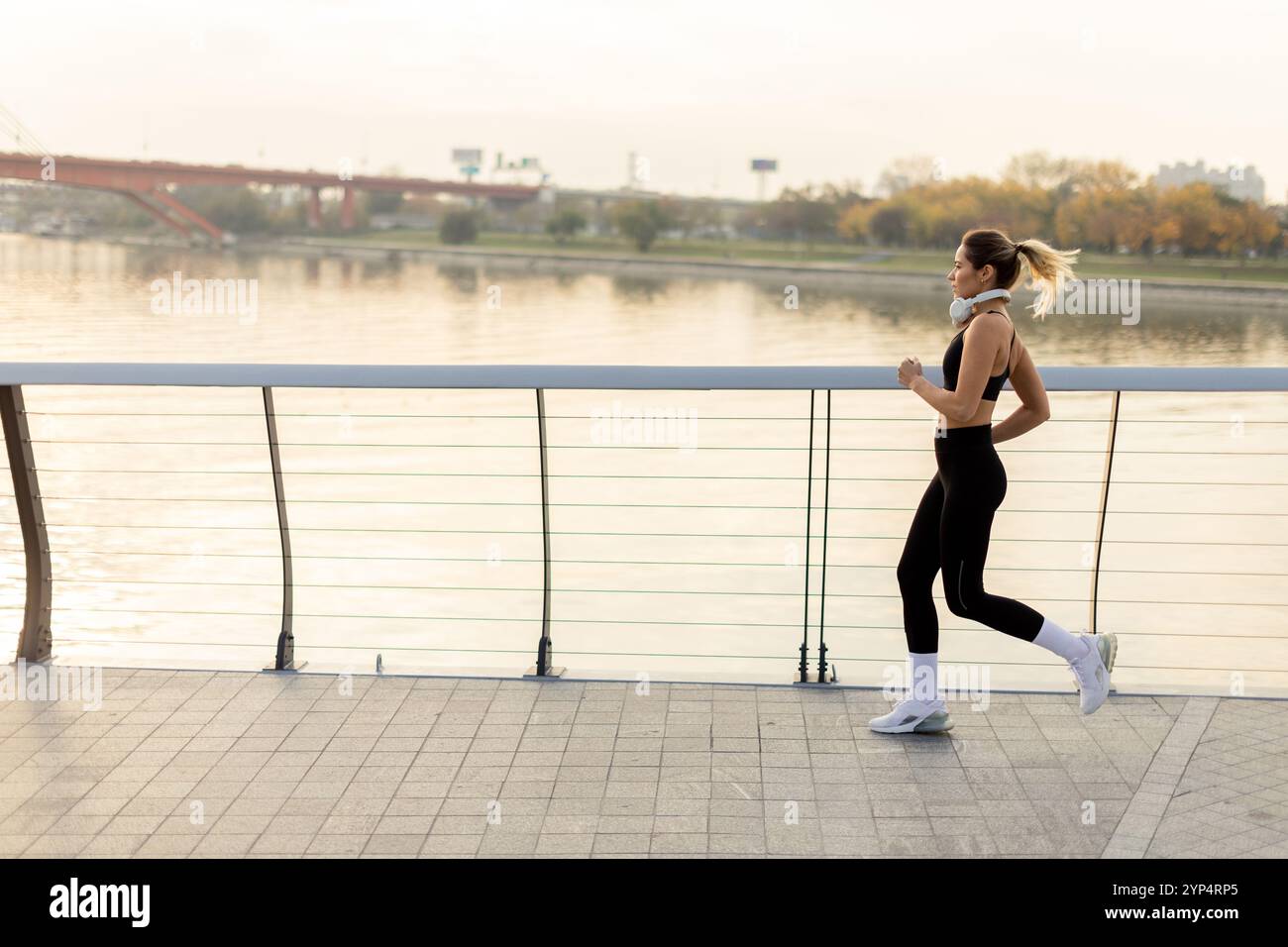 Un determinato corridore procede con sicurezza lungo un sentiero lungo il fiume, abbracciando l'aria frizzante del mattino, con lo skyline della città e gli alberi autunnali che offrono un'immagine Foto Stock