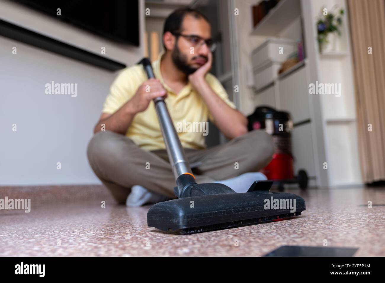 Un uomo siede sul pavimento in una casa moderna utilizzando un aspirapolvere. L'attenzione è rivolta alla pulizia e alle faccende domestiche, sottolineando la pulizia e la manutenzione domestica Foto Stock