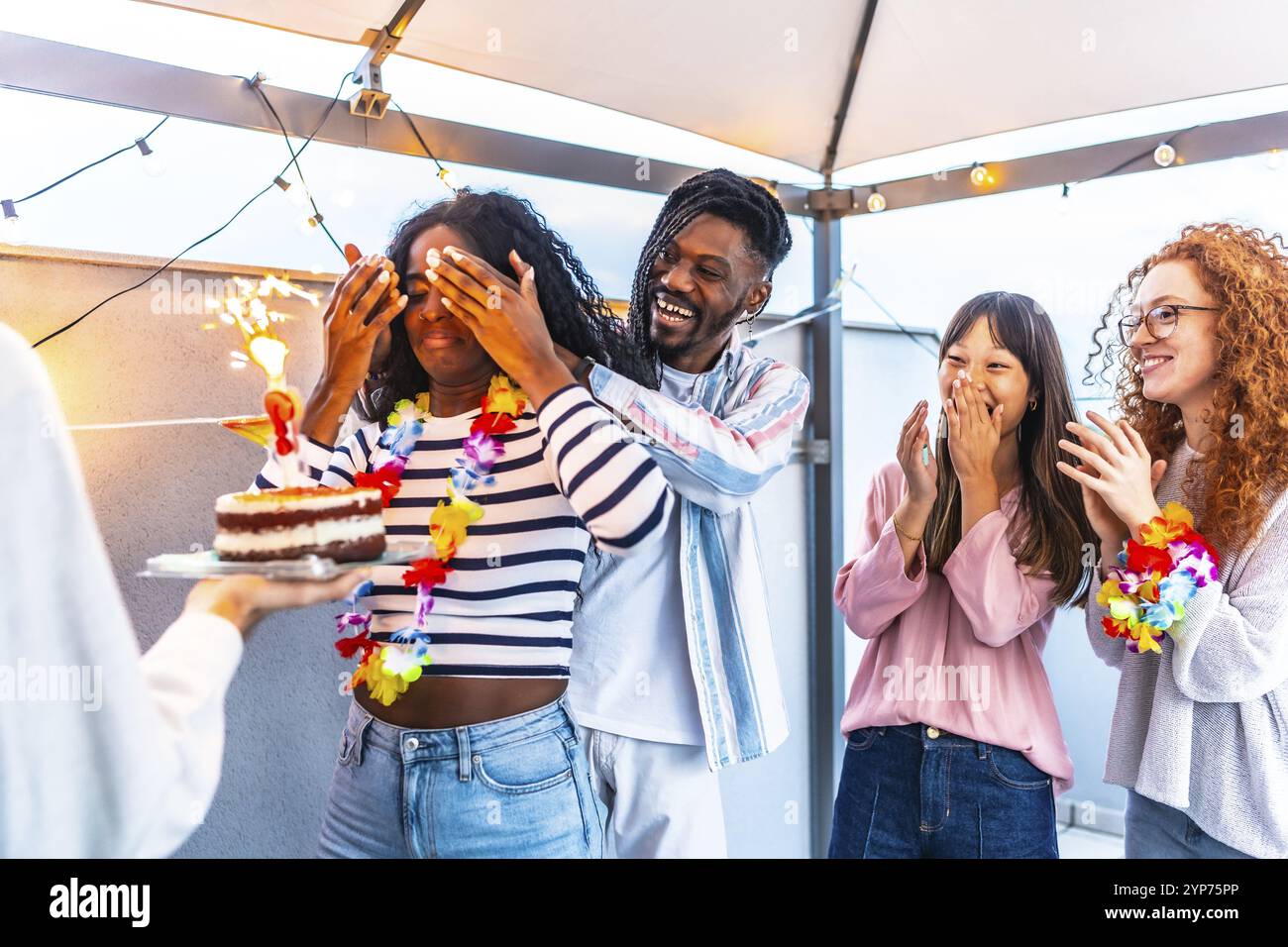 Giovane donna africana con gli occhi coperti che riceve una torta di compleanno da amici multietnici in terrazza Foto Stock